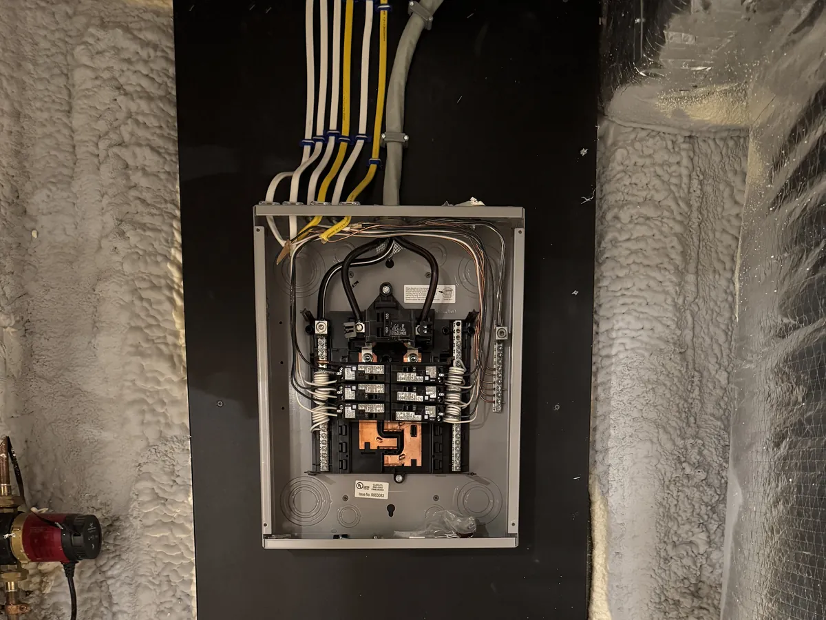 Open 100-amp residential sub-panel installation on a black backer board with spray-foam insulated walls, installed by A.J. LeBlanc Heating in Southern NH